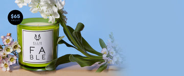 FABLE Candle between white and purple flowers with green leaves on a tan surface against a blue background with a black circle with blue text that reads "$65 Value"