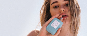 Woman with blonde hair holds SALT Eau de Parfum 50ml without cap against her chin with a gray background