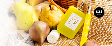 Sun Fruit 50ml Eau De Parfum without cap and Sun Fruit 10ml Eau De Parfum Travel Spray leaning on tan straw hat next to pears, a tan, brown, and white blanket, a cap, and a wicker basket with a black bubble with white text that reads "$33 value"