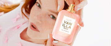 Woman with red hair and pale pink manicure wearing a pink shirt with bows on it holds VANILLA RICE Eau de Parfum 50ml without cap above her head on a pink background