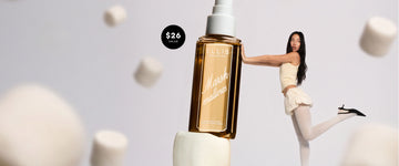 Model with brown hair in yellow tank top and skirt, white tights, and black heels faces to the side, stands on one leg and holds up with her arms a life size bottle of MARSHMALLOWS Perfume Mist without a cap stacked on top of a marshmallow with a black circle with white text that reads "$26 value" on a gray background with falling marshmallows