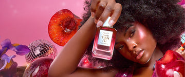 Woman with brown hair wearing a pink sequin tank top and pink highlighter holds PLUM TONIC Eau de Parfum 50ml against her head in one hand and a plum with a bite out of it in the other hand on a pink background with sliced plums, silver disco balls, and purple irises