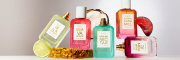Six 50mL perfume bottles from Ellis Brooklyn arranged on a white surface with decorative fruit including pineapple, plum, coconut, and lime against a light gray background. The bottles from left to right include Isla Sirena, Guava Granita, Miami Nectar, Queens Caramel, and Plum Tonic each with a white label and gold cap.