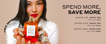 Model with brown hair wearing white jacket with pearls on sleeves holding Queens Caramel 50ml Eau De Parfum without cap and silver ornaments in two hands, on light brown background with black text that reads "Spend More, Save More Spend $75, Save 10% Use Code EB75 Spend $150, Save $15 Use Code EB150 Spend $250, Save 20% Use Code EB250"
