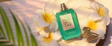 MIAMI NECTAR Eau de Parfum 50ml without cap lays on white cake with white and yellow flowers and gold eleven candles sitting on white textured blanket with green palm leaves in corner