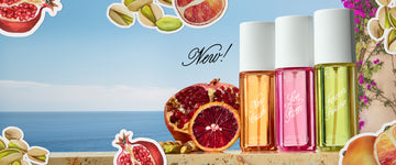 NEW CRUSH Perfume Mist 90ml, LOVE POM Perfume Mist 90ml, and FOREVER PISTACHIO Perfume Mist 90ml stand on tan marble next to a sliced pomegranate, a sliced blood orange, and pistachios in front of a tan wall with purple flowers, a blue ocean, and blue sky with drawn images of sliced pomegranate, sliced blood orange, and pistachios with a white outline with black text that reads "New!"