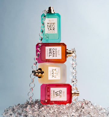 Miami Nectar 50ml Eau De Parfum without cap on top of Queens Caramel 50ml Eau De Parfum without cap on top of Vanilla Salt 50ml Eau De Parfum without cap on top of Guava Granita 50ml Eau De Parfum without cap, wrapped in beaded chain while resting on piled of beaded chain against blue background.