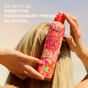 Femme aux cheveux blonds avec une bague en or et des bracelets tient le shampooing sec Amika x Ellis Brooklyn GUAVA GRANITA Perk Up contre l'arrière de sa tête, devant un ciel bleu et jaune avec un texte blanc qui dit « Ça sent le smoothie fraîchement pressé au soleil »