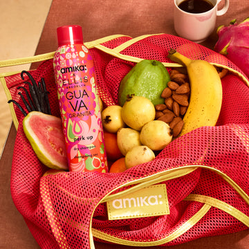 Shampoing sec Amika x Ellis Brooklyn GUAVA GRANITA Perk Up dans un sac en filet rose et jaune amika avec des goyaves, des gousses de vanille, des amandes, une banane et des oranges à côté d’un fruit du dragon et d’une tasse blanche avec une trace de rouge à lèvres rose remplie de café sur une table marron