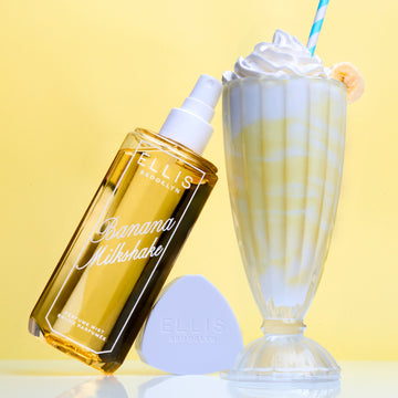 BANANA MILKSHAKE Perfume Mist 240ml without cap leans against cap and glass cup of yellow and white milkshake topped with whipped cream, a banana slice, and blue and white straw on a yellow and gray background 