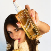 Model with brown hair wearing a yellow shirt looks up at the camera while eating a banana and holding BANANA MILKSHAKE 90ml perfume mist without a cap on a blue and white ombre background