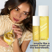 Model with brown hair and french manicure wearing yellow shirt and yellow earrings holding straw of glass with yellow smoothie and sliced bananas to mouth, on blue background with bottles of BANANA MILKSHAKE 240ml and 90ml and black text that reads "smells like a sweet treat on the boardwalk"