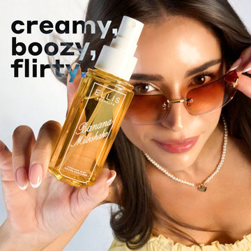 Model with brown hair wearing brown sunglasses, a yellow dress, and a pearl necklace looks up at the camera while holding BANANA MILKSHAKE 90ml perfume mist without a cap on a blue and white ombre background with black, white, and blue text that reads "creamy, boozy, flirty." 
