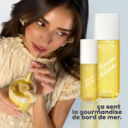 Modèle avec des cheveux bruns et une manucure française portant une chemise jaune et des boucles d'oreilles jaunes tenant une paille dans un verre avec un smoothie jaune et des bananes tranchées vers la bouche, sur un fond bleu avec des bouteilles de BANANA MILKSHAKE 240 ml et 90 ml et un texte noir qui dit 