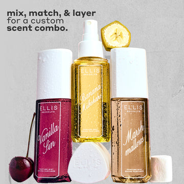VANILLA SIN Perfume Mist 240ml next to a cherry, BANANA Milkshake Perfume Mist 240ml sitting on top of it's cap, MARSHMALLOWS Perfume Mist 240ml with a slice on banana on top of it and a marshmallow next to it, all covered in water droplets on a gray, rough background with black text that reads "mix, match, and layer for a custom scent combo."
