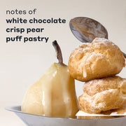 COCOA BLANCO fragrance notes displayed with a white chocolate-drizzled pear and two powdered puff pastries on a silver tray, with a spoon behind them and black text that reads "notes of white chocolate, crisp pear, puff pastry" on a cream background.