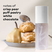COCOA BLANCO perfume mist next to a pear, choux pastry, and spoon in a silver plate on a light beige background with text reading 'notes of crisp pear, puff pastry, white chocolate'.