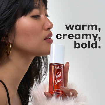 Woman with black hair and bangs wearing gold hoop earrings, a black tank top, and fuzzy, white sweater holds FIRST DATE Perfume Mist 90ml against her chin with black text that reads "warm, creamy, bold." on a white background