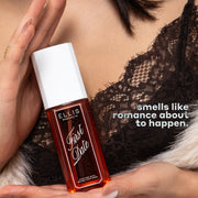 Model with black hair in black lace top and white fuzzy sweater holds FIRST DATE Perfume Mist 90ml between her two palms in front of her chest with white text that reads "smells like romance about to happen."