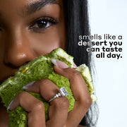Woman with black hair wearing a diamond ring and holding green cake with white icing against a white background with black and gray text that reads "smells like a dessert you can taste all day."