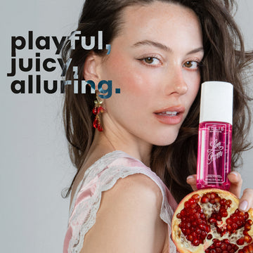 Model with brown hair wearing pink silk and white lace tank top and pink earrings holds LOVE POM Perfume Mist 90ml and half of a pomegranate against a gray background with black and gray text that reads "playful, juicy, alluring."