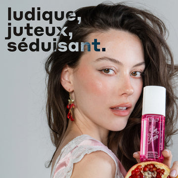 Un modèle aux cheveux bruns portant un débardeur en soie rose et dentelle blanche et des boucles d'oreilles roses tient le parfum LOVE POM Perfume Mist 90 ml et une moitié de grenade devant un fond gris avec un texte noir et gris qui dit « ludique, juteux, séduisant ».