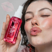 Femme aux cheveux bruns portant un débardeur en soie rose et dentelle blanche et des boucles d'oreilles roses, faisant une moue et tenant une bouteille de LOVE POM Perfume Mist 90ml sans bouchon sur un fond gris avec un léger contour de rouge à lèvres rose