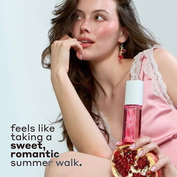 Woman with brown hair wearing pink silk and white lace tank top and pink earrings holds LOVE POM Perfume Mist 90ml on half of a pomegranate and places hand against her lips against a gray background with black text that reads "feels like taking a sweet, romantic summer walk."