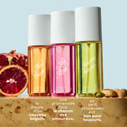 Brume de parfum NEW CRUSH 90ml, Brume de parfum LOVE POM 90ml et Brume de parfum FOREVER PISTACHIO 90ml sur du marbre beige entre des tranches de grenade, d’orange sanguine et des pistaches, sur un fond bleu clair avec un texte blanc qui dit « la magie d’un nouveau béguin. Une promenade dans le chemin des amoureux. ça sent délicieusement bon pour toujours. »