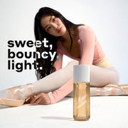 Model with brown hair wearing a pink top, yellow leotard, white tights, and pink ballet slippers sits and stretches forward with a MARSHMALLOWS Perfume Mist 240ml sitting on the white floor in front of her on a white background with white, black, and blue text that reads "sweet, bouncy, light."