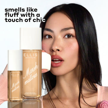 Model with brown hair wearing a peach top with a pink rose eats a marshmallow with MARSHMALLOWS Perfume Mist 240ml and 90ml in front of her on a white background with black text that reads "smells like fluff with a touch of chic"