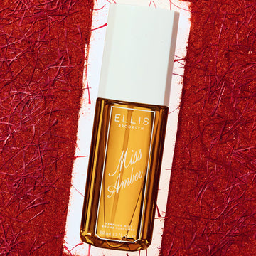 Miss Amber perfume mist 90mL bottle with an amber glass body and a white cap, set against a textured red and white background.