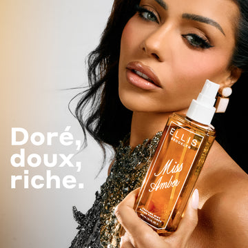 Une femme aux cheveux foncés et portant un haut à perles tient un flacon de 90 mL de brume parfumée Miss Amber devant un fond beige chaud, avec le texte « doré, doux, riche. »