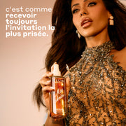 Une femme portant une robe transparente ornée de paillettes tient un flacon de 90 mL de parfum en brume Miss Amber devant un fond beige chaud, avec un texte promotionnel à gauche qui dit « C'est comme recevoir toujours l'invitation la plus prisee. »