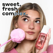 Woman with blonde hair in a pink sweater and gold hoop earrings looks up and holds MISS STRAWBERRY Perfume Mist 90ml against one cheek and a pink pen with a pink pompom on the end against the other cheek with black and blue text that reads "sweet, fresh, complex." all on a white background.
