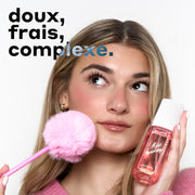 Femme aux cheveux blonds portant un pull rose et des boucles d'oreilles créoles dorées, regardant vers le haut et tenant le parfum MISS STRAWBERRY Perfume Mist 90ml contre une joue et un stylo rose avec un pompon rose au bout contre l'autre joue, avec un texte noir et bleu qui dit « doux, frais, complexe. » le tout sur un fond blanc.