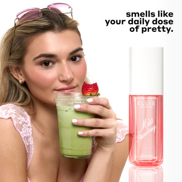 Woman with blonde hair wearing a pink lace tank top, gold hoop earrings, and pink sunglasses on her head holds a clear plastic cup of matcha tea with a half eaten strawberry on top of it next to MISS STRAWBERRY Perfume Mist 90ml with black text that reads "smells like your daily dose of pretty." on a white background