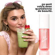 Femme aux cheveux blonds portant un débardeur en dentelle rose, des boucles d'oreilles créoles en or et des lunettes de soleil roses sur la tête, tenant un gobelet en plastique transparent de thé matcha avec une fraise à moitié mangée dessus à côté du parfum en brume MISS STRAWBERRY 90 ml avec un texte noir qui dit « ca sent votre dose quotidienne de beauté » sur fond blanc.