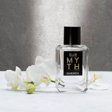 Myth 50ml Eau de Parfum sur une table blanche à côté de fleurs blanches contre un mur gris.