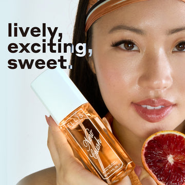 Model with black hair wearing orange tank top and headband holds NEW CRUSH Perfume Mist 90ml and slice of blood orange against white background with black and white text that reads "lively, exciting, sweet."