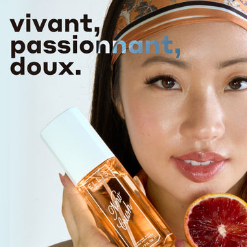 Un mannequin aux cheveux noirs portant un débardeur orange et un bandeau tient le parfum en brume NEW CRUSH 90 ml et une tranche d'orange sanguine sur fond blanc avec un texte en noir et blanc qui dit « vivant, passionnant, doux. »