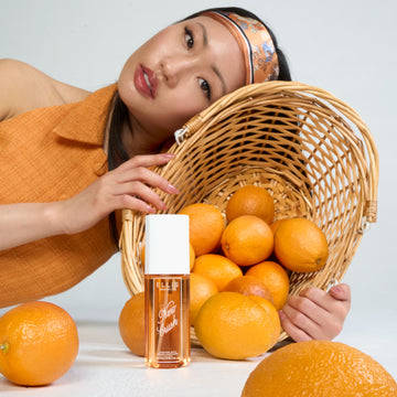 Femme aux cheveux noirs portant un bandeau et un débardeur orange et bleu, appuyée contre un panier rempli d'oranges derrière une bouteille de parfum en brume NEW CRUSH 90 ml et des oranges sur fond blanc