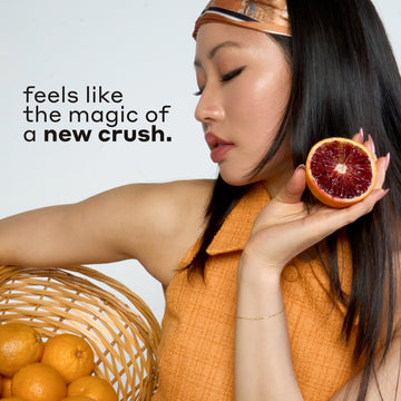 Woman with black hair wearing an orange and blue headband and tank top holds tan basket filled with oranges and a sliced blood orange against white background with black text that reads "feels like the magic of a new crush."