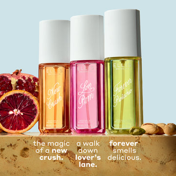 NEW CRUSH Perfume Mist 90ml, LOVE POM Perfume Mist 90ml, and FOREVER PISTACHIO Perfume Mist 90ml on tan marble between sliced pomegranate, blood orange, and pistachios against a light blue background with white text that reads "the new magic of a new crush. a walk down lover's lane. forever smells delicious."
