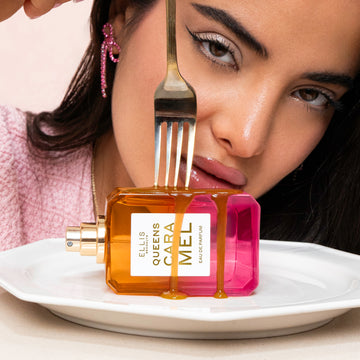 Model with black hair wearing pink diamond earring, gold necklace, and pink shirt holding fork to Queens Caramel 50ml Eau De Parfum bottle without cap, covered in melted caramel on white plate, on white table, on white background.