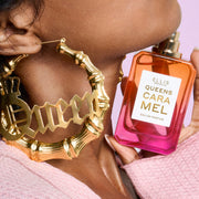Close-up of woman wearing a pink sweater and gold hoop earrings that say "Queen" holding QUEENS CARAMEL Eau de Parfum 50ml without cap under her chin on pink background