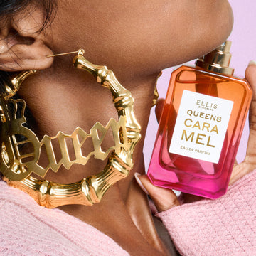 Close-up of woman wearing a pink sweater and gold hoop earrings that say "Queen" holding QUEENS CARAMEL Eau de Parfum 50ml without cap under her chin on pink background