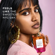 Woman with black hair wearing a pink sweater and gold hoop earrings that say "Queen" holds QUEENS CARAMEL Eau de Parfum 50ml without cap under her chin on pink background with white text that reads "Feels like the sweetest NYC day."