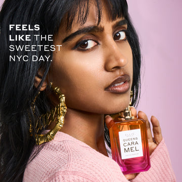 Woman with black hair wearing a pink sweater and gold hoop earrings that say "Queen" holds QUEENS CARAMEL Eau de Parfum 50ml without cap under her chin on pink background with white text that reads "Feels like the sweetest NYC day."