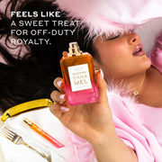 Model with black hair and French manicure wearing gold necklace and pink satin robe with feathers at cuffs laying down on pink couch next to gold tray with napkin, fork, and Queens Caramel 10ml Eau De Parfum Travel Spray, holding Queens Caramel 50ml Eau De Parfum without cap to camera, with white text that reads 